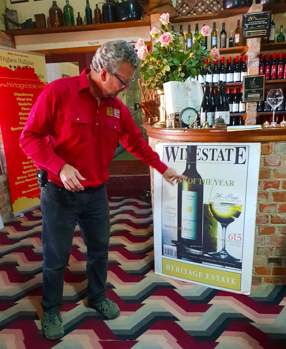 Image of a man in red shirt and dark trousers pointing to a large poster attached to a bar. The poster is of a bottle of wine next to a glass filled with wine