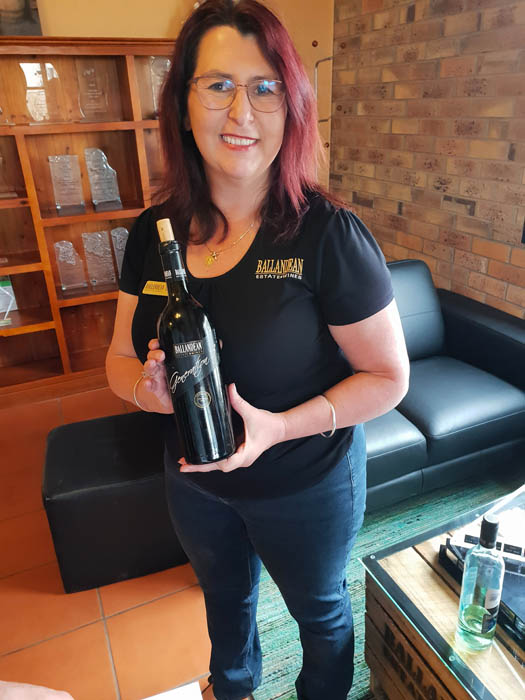 Image of a smiling woman wearing blue jeans and black tshirt holding a corked bottle of wine