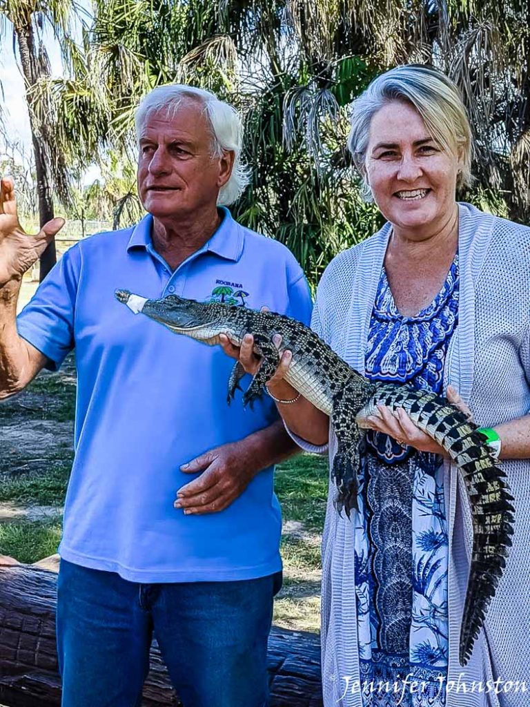 An older man wearing blue shirt stands next to woman wearing dress and cardigan who is holding a baby crocodile
