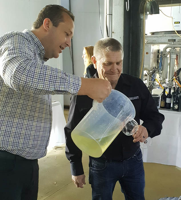 Image of two men standing next to each other one if pouring wine from a large plastic jug into the other man's wine glass