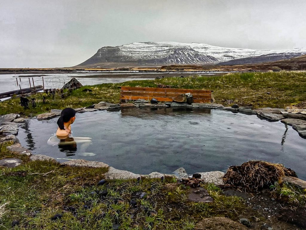 Woman sits in open air hot pool looking out over the ocean towards a large mountain