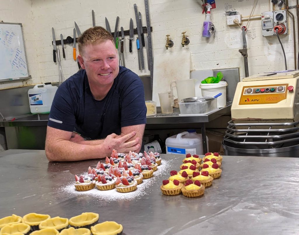 Image of a man leaning on a steel table near a collection of homemade pastries