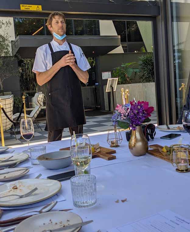 Image of a man standing in front of a long table wearng an apron on top of his clothes signifying he is a chef
