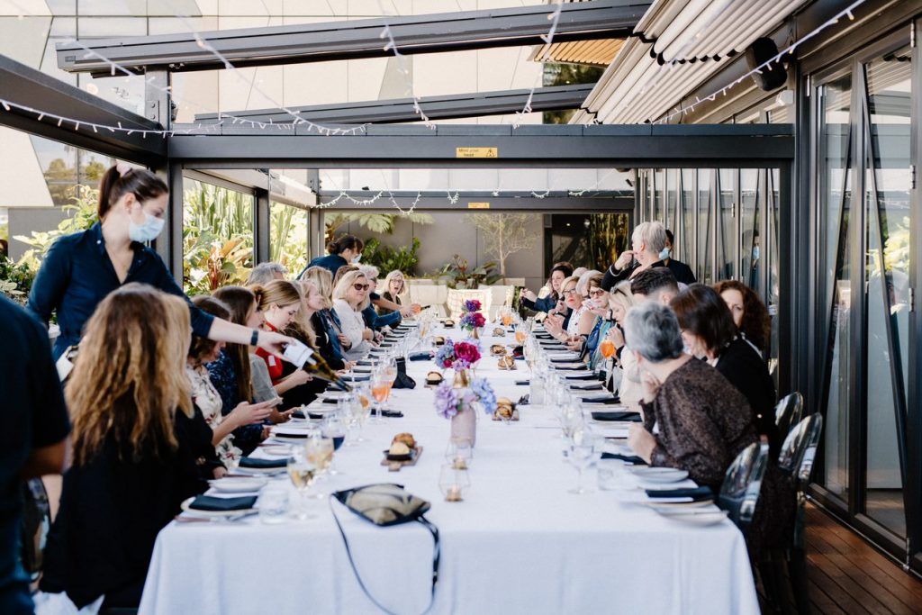Image of a large long rectangular shaped table with people seated around the table eating lunch and drinking wine