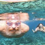 IMage taken underwater of a man swimmng wearing goggles and a young girl next to him also wearin goggles