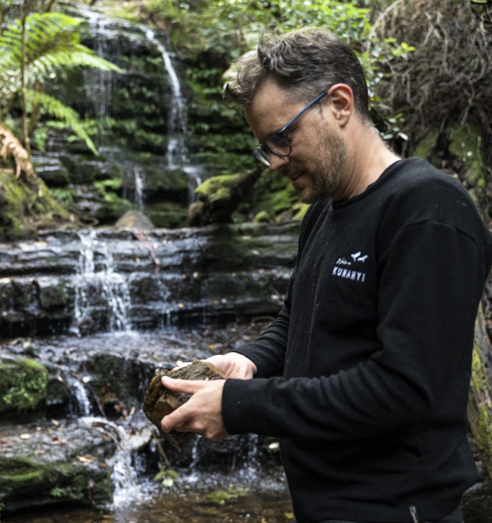 Image of a man wearing a black shirt holding a piece of rock a waterfall is in the background
