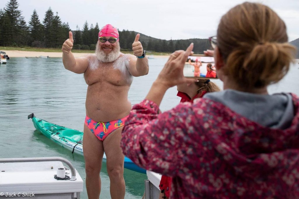 Image of a man standing in his swimmimg trunks with swim goggles and bathing cap holding his thumbs up to a woman taking his photo