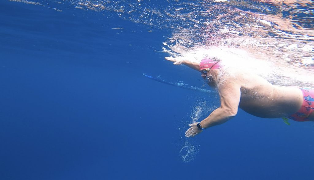 Image taken underwater of a man swimming in the ocean he is wearing pink swimming trunks and a pink bathing cap