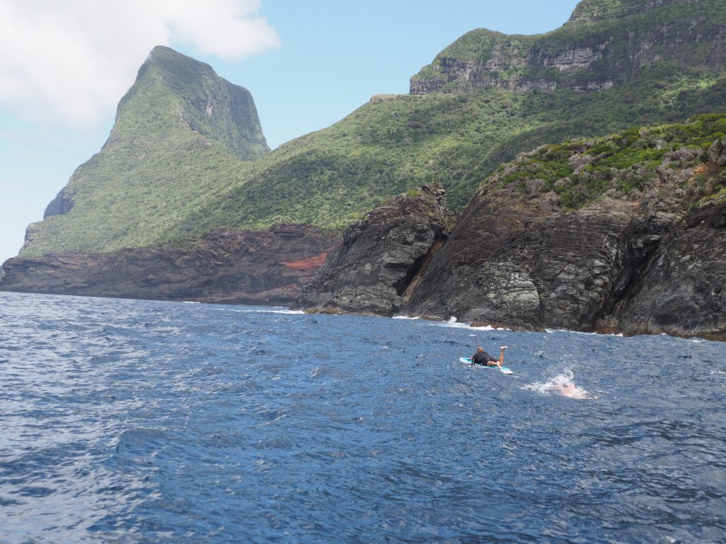 Image of a man on a lying down on a paddle board with a man swimming behind in the open ocean large mountain in the background