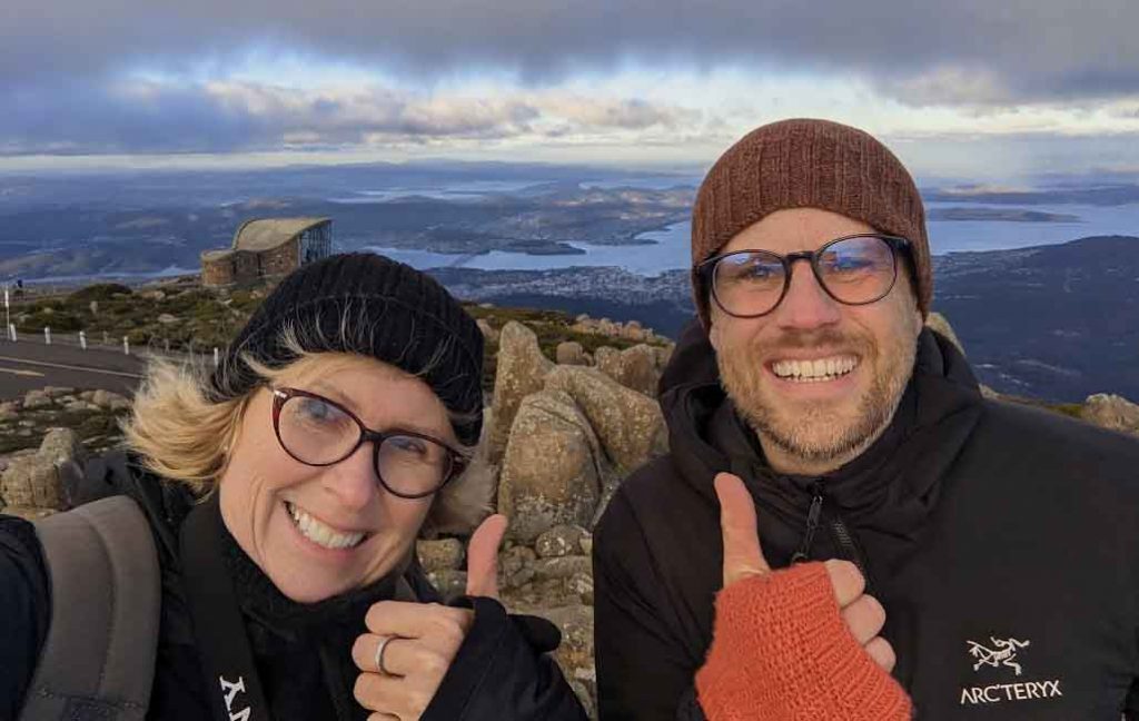 Two people wearing beanies and warm coats standing on top of a mountain with thumbs up