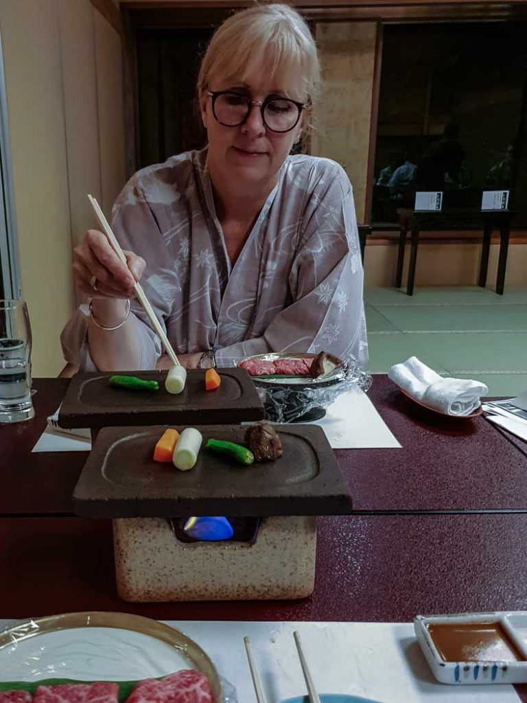 Image of a woman sitting at a table wearing a kimino and using chopsticks to pick up food from plates on the table