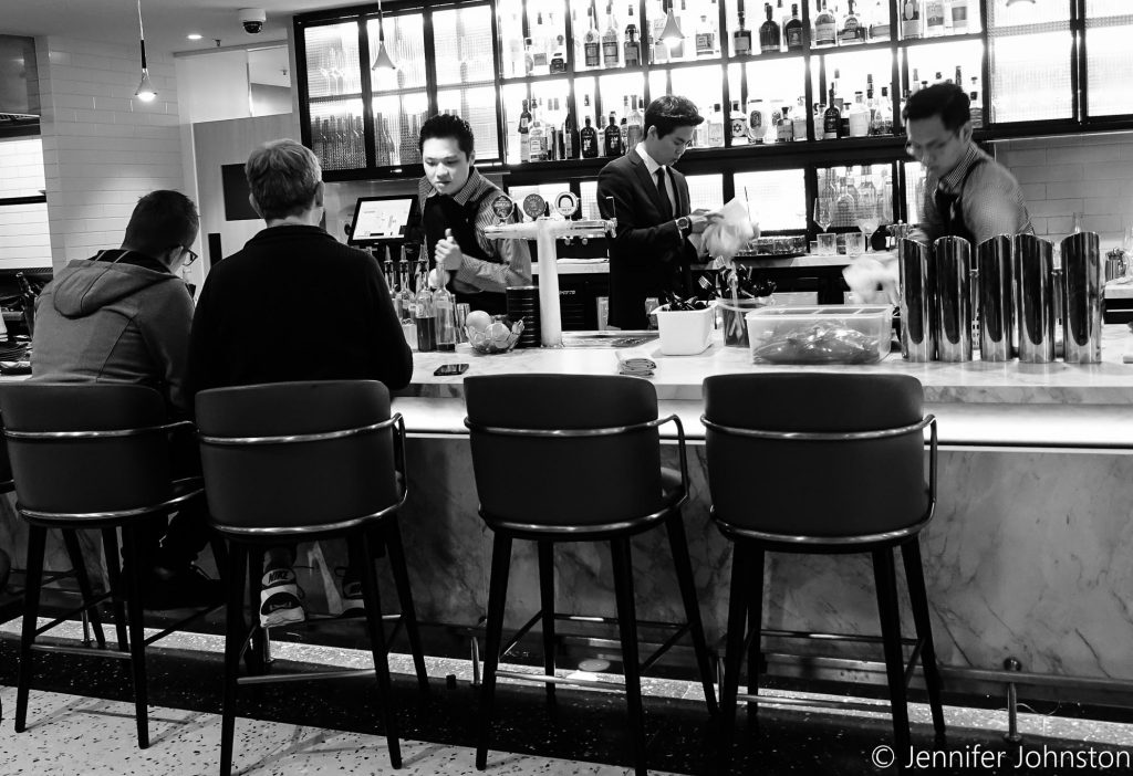 Image in black and white of a bar and people sitting on high stools on one side of the bar and bar staff making drinks on the other sideone side