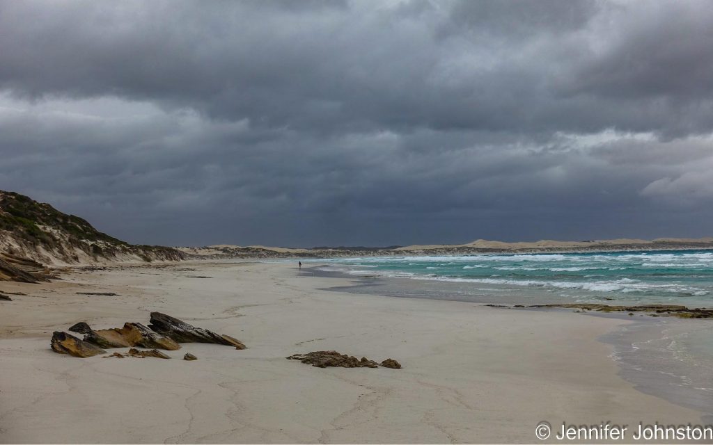 Image of a long white sandy beach with one person in the distance the sea is rough looking and the sky is stormy