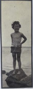 Black and white image of a young child wearing swimming trunks standing on a rock