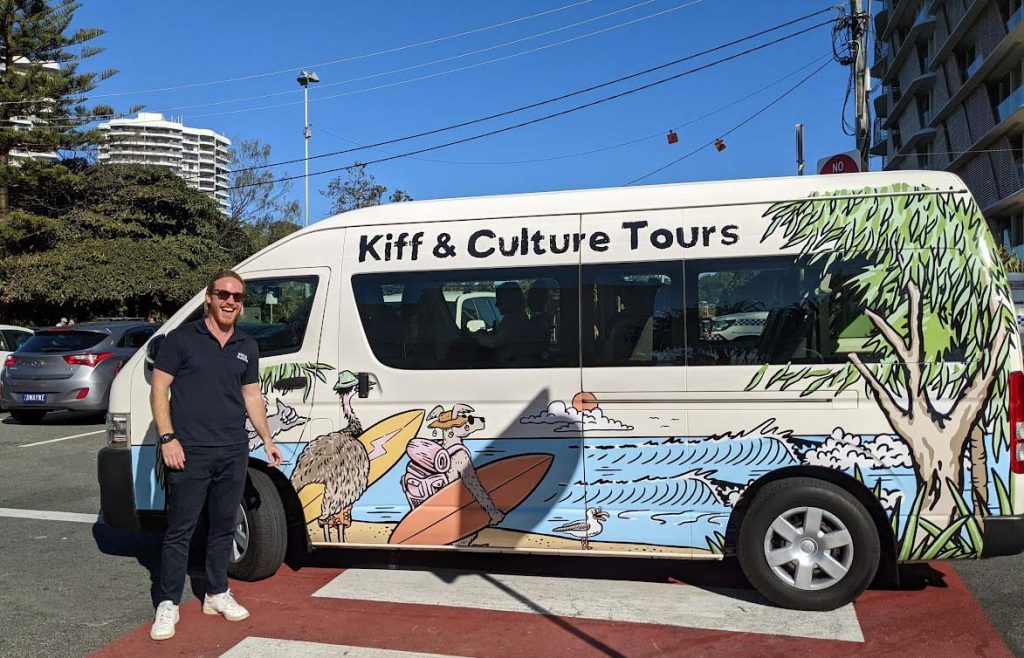 Image of a man wearing black trousers and shirt standing in front of a bus with the words Kiff and Culture on the side