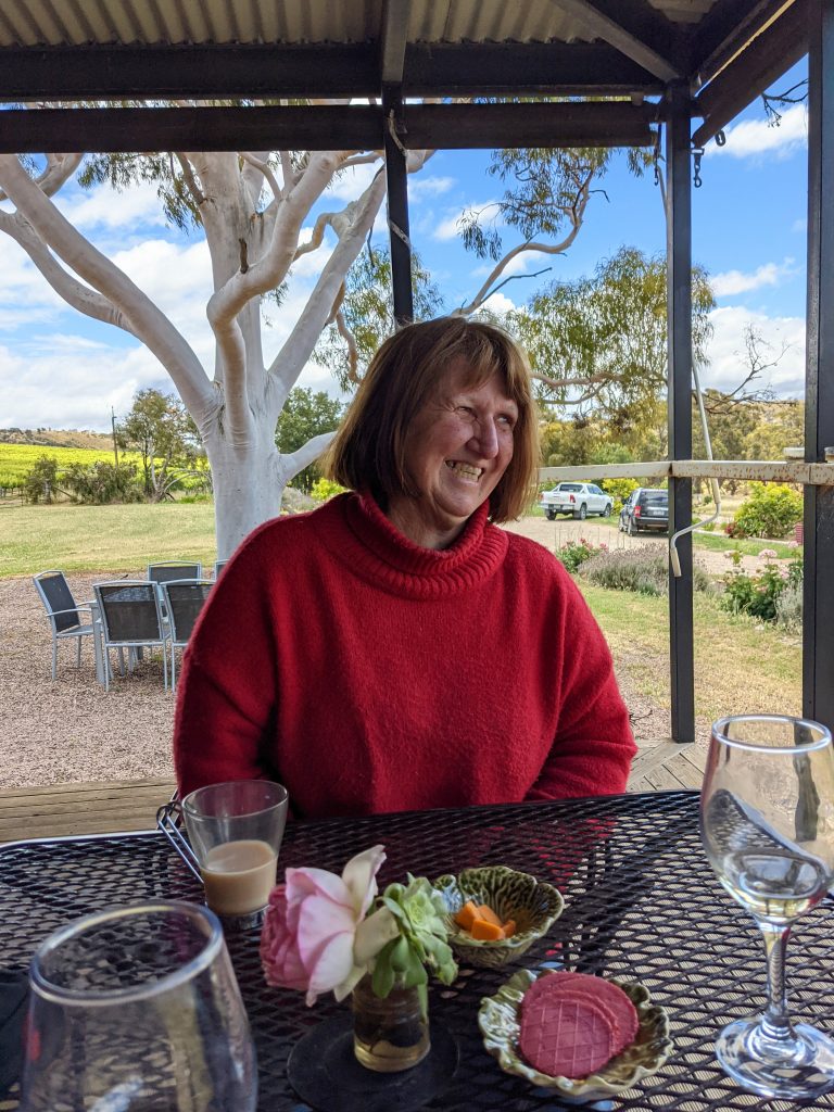 Image of a smiling woman wearing a red jumper sitting at a table with a glass of wine a cup of coffee on the table