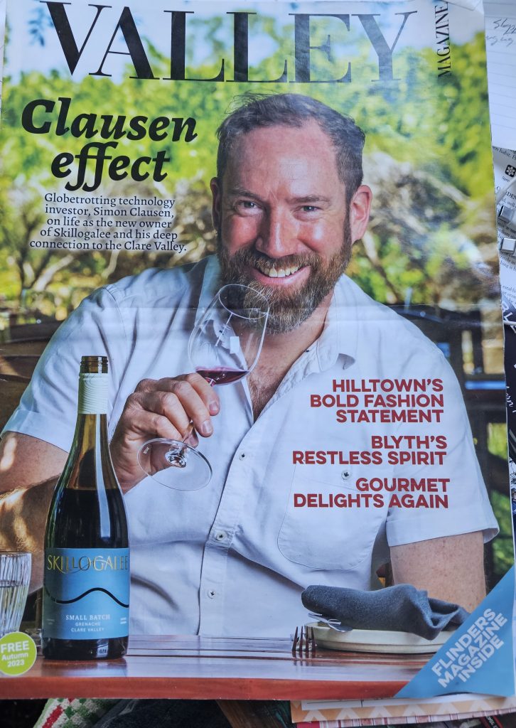 Image of a front cover of a magazine showing a man sitting down holding a glass of wine with a bottle of wine on the table