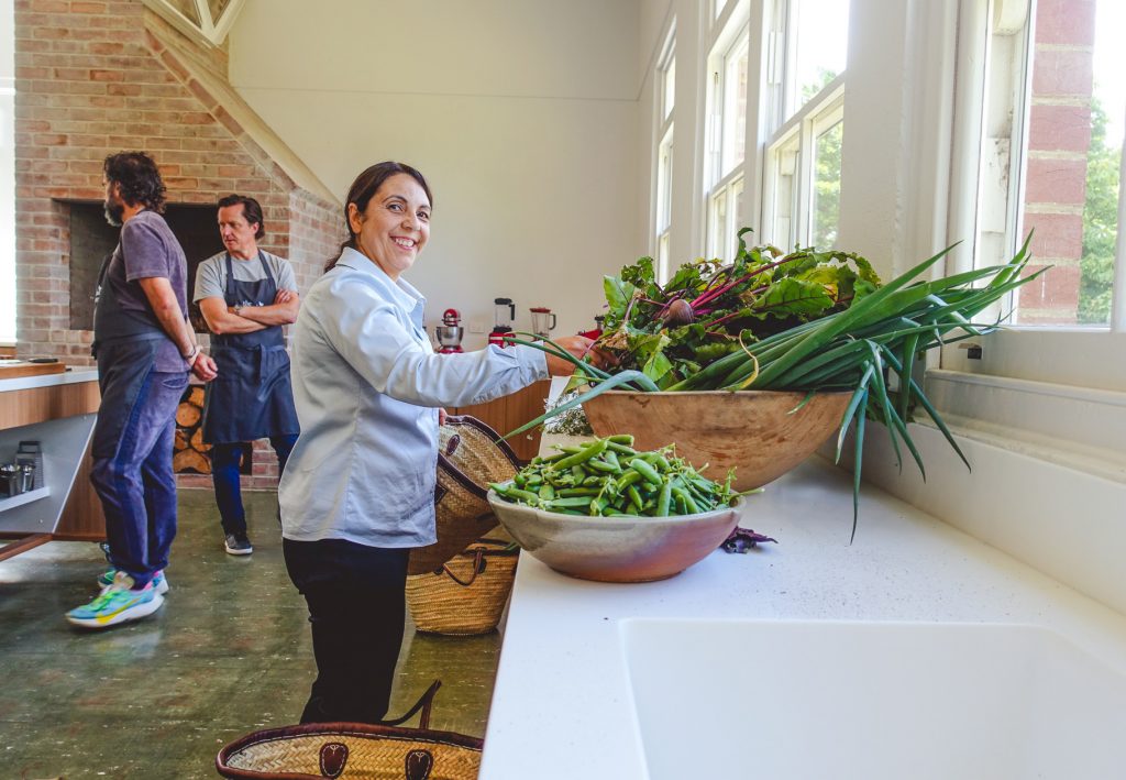 Image of a smiling woman standing at a kitchen bench that has a large wooden bowl filled with fresh garden salad and vegetables