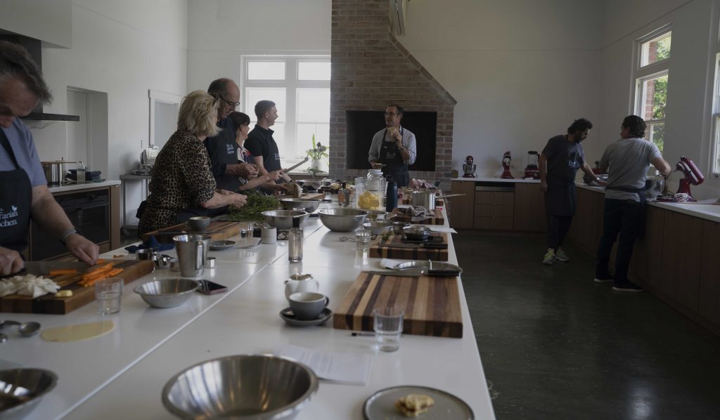 Image of a commercial kitchen and people standing on either side of a long kitchen bench receiving instruction
