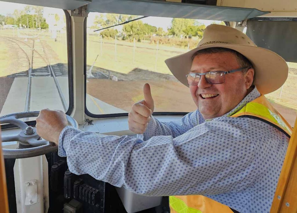 Image of a man wearing an old Akubra hat and a high vis vest sits in the drivers seat of a train