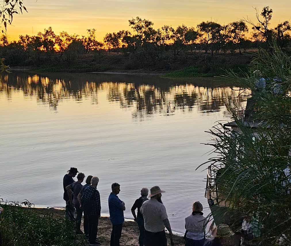 Image of a group of people standing on a river bank watching the sunset