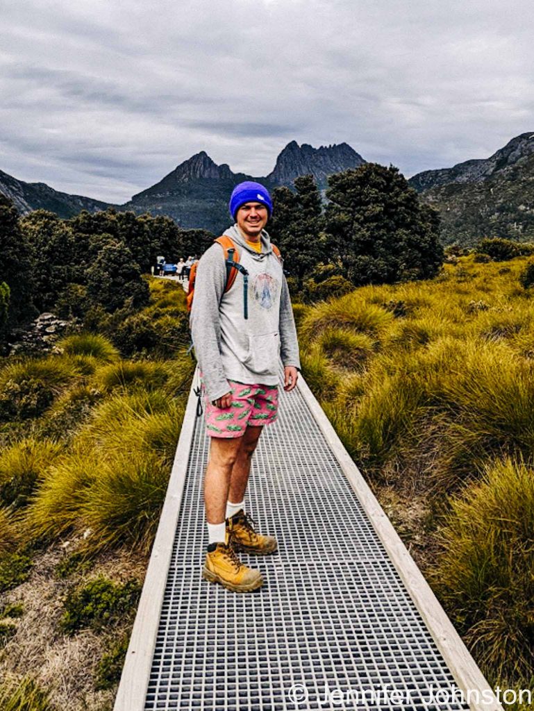 Image of a young main dressed in shorts and a long sleeved hoodie with and a purple beanie standing on the walking trail with the outline of a mountain behind him