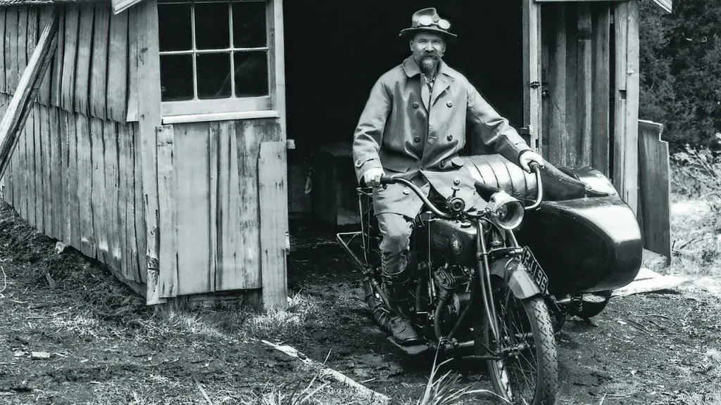 Black and White image of a man sitting on a motorbike he is wearing an overcoat, trousers and boots. On his head is a crumpled hat with goggles