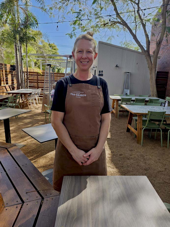 Image of a woman wearing a black apron with the words Branch Cafe. She is standing in a courtyard of a restaurant she is beside a table i